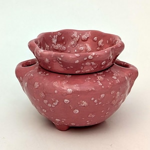 Op de afbeelding: Een tweedelige, handgemaakte keramische pottenbakkerset in een stoffige roze kleur met witte spikkels. Het onderste deel is een ronde vaas met twee handvatten en drie kleine voetjes. Het bovenste deel is een kleinere, geschulpte kom.