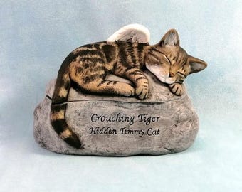 May include: A ceramic figurine of a tabby cat with angel wings, sleeping on a rock. The rock has the inscription "Crouching Tiger Hidden Timmy Cat".