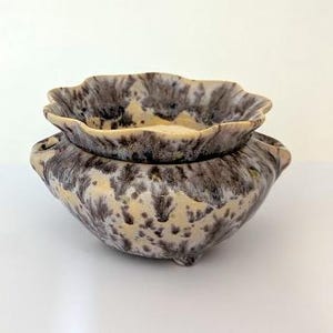 May include: A ceramic planter with a scalloped rim and two side handles. The vessel features a cream-coloured base glaze with a mottled, dark grey and black drip pattern that covers the exterior and the interior rim, resting on small feet.