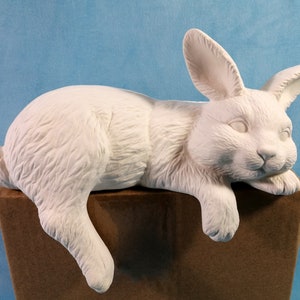 May include: White ceramic bunny figurine lying down with its head resting on its paws. The bunny is on a brown surface.