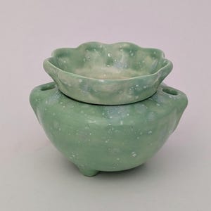 Op de afbeelding: Een tweedelige keramische set in een zachte groene tint met witte spikkels. Het onderste deel is een afgeronde kom met drie kleine voetjes en twee handvatten. Het bovenste deel is een kleinere kom met geschulpte rand.