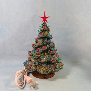 May include: A ceramic Christmas tree with a red star on top. The tree is green with colorful lights and sits on a brown base. A white electrical cord and plug are visible.