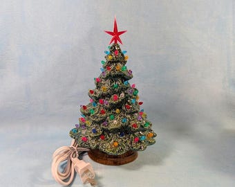 May include: A ceramic Christmas tree with a red star on top. The tree is green with colorful lights and sits on a brown base. A white electrical cord and plug are visible.