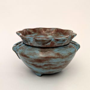 May include: A two-piece ceramic bowl set with a distressed blue and brown finish. The top bowl has a scalloped edge and sits atop a larger, rounded bowl with small feet and handles. The set has a rustic, handcrafted appearance.