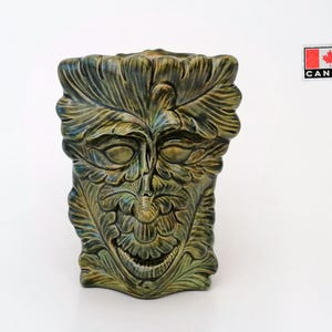 Puede incluir: Una escultura de cerámica verde de una cara con detalles de hojas. La cara tiene una boca ancha y los ojos cerrados. La escultura es texturizada y tiene un aspecto rústico.