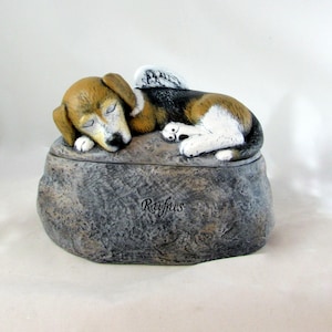 May include: A ceramic pet urn shaped like a rock with a sleeping dog figurine on top. The dog is brown, black, and white with angel wings. The urn is inscribed with the name "Rufus".