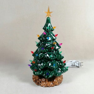 May include: A ceramic Christmas tree with a glossy green finish. The tree is adorned with colorful lights and topped with a gold star. The base is brown and textured. A white power cord with a plug is attached.