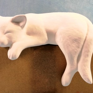 Op de afbeelding: Witte keramische kattenfiguur in een slaaphouding. De kat ligt op zijn zij met zijn kop op een bruine doos.
