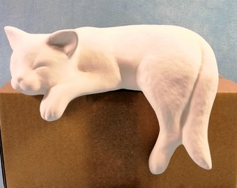May include: White ceramic cat figurine in a sleeping pose. The cat is lying on its side with its head resting on a brown box.