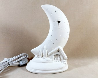 May include: A white ceramic crescent moon lamp featuring a cut-out star design and a small nativity scene at the base. The lamp includes a white electrical cord with a switch, showcasing a snowy landscape with trees and a stable structure.