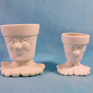 May include: Two white ceramic planter pots shaped like faces with feet. The larger pot has a closed-eye expression, while the smaller pot has an open-mouthed smile.