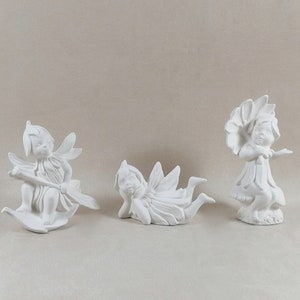 May include: Three white ceramic fairy figurines. One fairy is sitting on a leaf holding a stick, another is lying on its stomach with wings spread out, and the third is standing with a flower on its head.