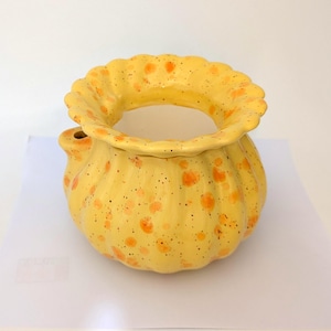 May include: A yellow ceramic vase with a frilled rim and a rounded body. The vase is speckled with orange spots and has a small spout. The vase is on a white surface.