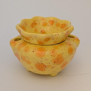 May include: A yellow ceramic oil warmer with a scalloped top dish and a rounded base featuring three small feet. The surface is decorated with orange spots and dark brown speckles throughout. Two small circular openings are visible on the sides.
