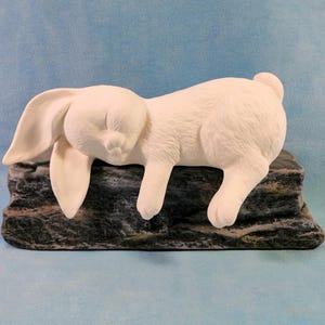 Może przedstawiać: Biała ceramiczna figurka królika śpiącego na szarej i czarnej podstawie skalnej. Królik ma długie, zwisające ucho i leży na boku z głową opartą o skałę.