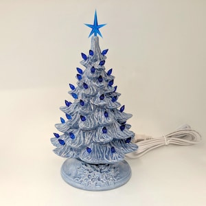 Könnte beinhalten: Ein Keramik-Weihnachtsbaum mit hellblauer Glasur, der mit zahlreichen kleinen blauen Kunststoffbirnen in den Zweigen und einem blauen Stern als Spitze versehen ist. Der Sockel ist mit einem geformten Stechpalmenblattmuster verziert.