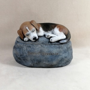 May include: A ceramic urn shaped like a rock with a sleeping beagle dog on top. The urn is inscribed with the name "Jason P. Baudanza".