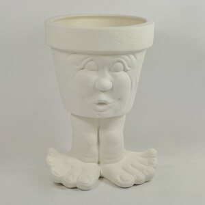 May include: A white ceramic planter shaped like a head with a wide rim and a face featuring closed eyes, a nose, and lips. The planter is supported by two feet with visible toes at the base, creating a whimsical character design in a matte finish.