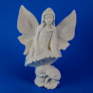 Peut inclure: Une figurine de fée en céramique blanche assise sur un champignon avec les ailes déployées. La fée porte une couronne sur la tête et une robe.
