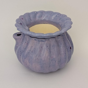 Peut inclure: Pot en céramique lavande, de forme bulbeuse et à finition mouchetée. Il présente un bord festonné et un petit bec. L'intérieur est de couleur crème. Ce pot décoratif pourrait servir de jardinière ou de petit récipient.