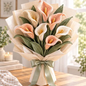 May include: A bouquet of crocheted calla lilies in shades of soft pink and cream, featuring yellow spadices and dark green leaves. The flowers are wrapped in light beige paper and tied with a sage green satin ribbon bow at the base.