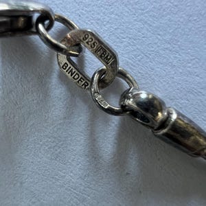 Könnte beinhalten: Ein silbernes Kettenglied mit einem rechteckigen Anhänger, der mit dem Text 925 FBM und BINDER geprägt ist. Das metallische Glied ist mit einem abgerundeten silbernen Element verbunden und zeigt deutliche Punzen auf der polierten Oberfläche.