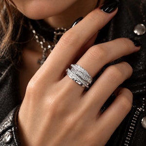 Puede incluir: Un anillo de varias bandas en tono plateado con una fila central de piedras de corte baguette flanqueada por dos filas de piedras de corte brillante redondo. Las bandas están interconectadas en un diseño cruzado.