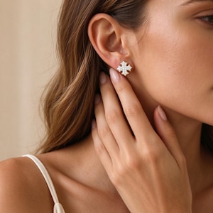 Puede incluir: Un pendiente de botón en forma de cruz, con un borde de metal texturizado y un centro relleno de pequeñas piedras claras y brillantes. El pendiente se lleva en el lóbulo de la oreja, mostrando un diseño detallado y ornamentado.