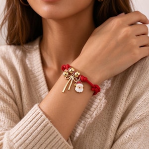 Puede incluir: Una pulsera de cordón rojo con cuentas de metal dorado, un dije de lazo dorado y un pequeño dije de esmalte con forma de Papá Noel. La pulsera se asegura con un cierre de nudo y presenta elementos decorativos de temática navideña.