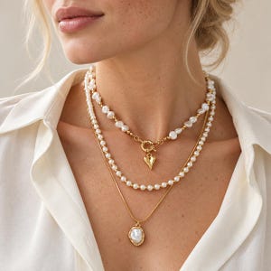 Puede incluir: Un conjunto de collares superpuestos que presenta una hilera superior de perlas blancas irregulares con un cierre de palanca dorado y un pequeño dije de corazón, una hilera media de perlas blancas uniformes y una cadena dorada con colgante.