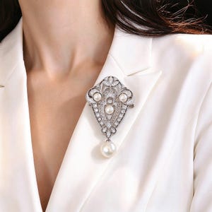 Puede incluir: Un broche de tono plateado con un intrincado diseño de filigrana adornado con múltiples piedras claras pequeñas y tres perlas blancas redondas engastadas en el marco, con una perla blanca más grande colgando del centro.