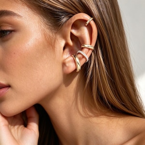 Puede incluir: Un conjunto de cuatro pendientes tipo ear cuff dorados colocados en el cartílago y la concha de la oreja. Tres tienen un acabado metálico liso y pulido, mientras que el tercero desde arriba está incrustado con una fila de piedras claras.