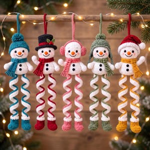 Könnte beinhalten: Fünf gehäkelte Schneemann-Ornamente, die in einer Reihe hängen. Jeder Schneemann hat einen weißen Körper, eine karottenförmige Nase und lange, spiralförmige Beine in abwechselnden Farben. Sie tragen verschiedene Mützen, Schals und Ohrenschützer.