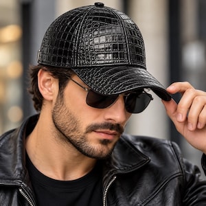 Puede incluir: Una gorra de béisbol negra con un patrón de piel de cocodrilo brillante y texturizado. La gorra tiene una corona estructurada, una visera curva y un pequeño botón en la parte superior central. El material tiene un acabado reflectante.