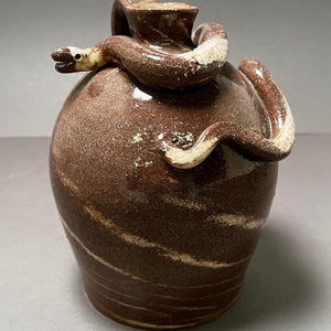 May include: A brown ceramic vase with a snake wrapped around it. The snake is a lighter brown with white accents. The vase has a round body and a small opening at the top. The snake's head is near the top of the vase.