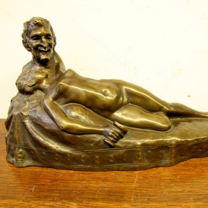 May include: Bronze sculpture of a reclining nude figure with a man standing over them. The sculpture is on a base that resembles a rug.