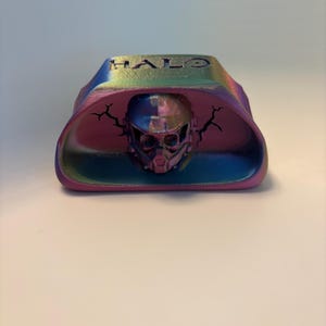 May include: A 3D printed iridescent display stand featuring the word HALO embossed on the top surface. The center of the stand contains a detailed helmet sculpture flanked by two black lightning bolt graphics on the curved inner wall.