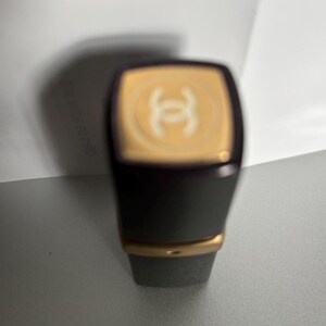 May include: A dark gray lipstick tube with a gold band and a gold top featuring a white logo. The lipstick has a rectangular shape with rounded corners. The background is a light gray surface.