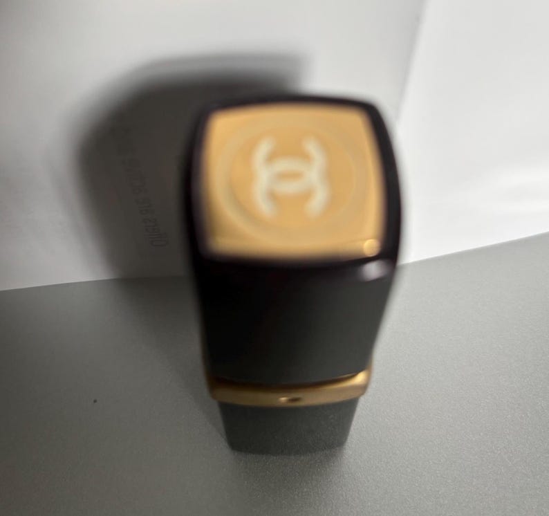 May include: A dark gray lipstick tube with a gold band and a gold top featuring a white logo. The lipstick has a rectangular shape with rounded corners. The background is a light gray surface.