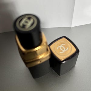 May include: Black lipstick tube with gold accents and a matching cap. The cap and the top of the tube feature a white logo. The lipstick is partially unscrewed, revealing the product.