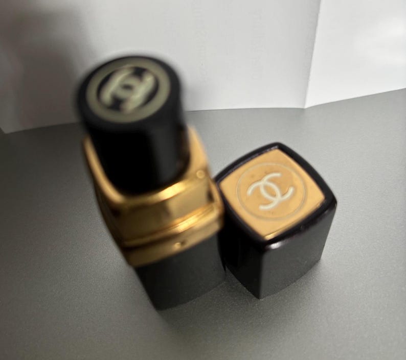 May include: Black lipstick tube with gold accents and a matching cap. The cap and the top of the tube feature a white logo. The lipstick is partially unscrewed, revealing the product.
