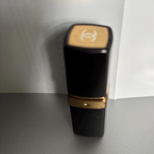 May include: A black lipstick tube with a gold band and a beige top featuring a white logo. The lipstick is on a gray surface with a white background.