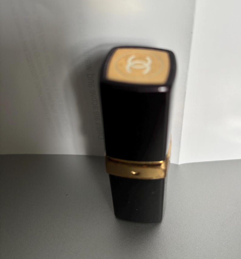 May include: A black lipstick tube with a gold band and a beige top featuring a white logo. The lipstick is on a gray surface with a white background.