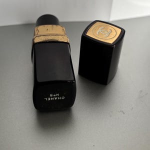 May include: A black perfume bottle with a gold-colored rim and a matching cap. The cap features a gold emblem. The bottle is labeled "CHANEL N&deg;5" in white text. The bottle is rectangular and has a classic design.