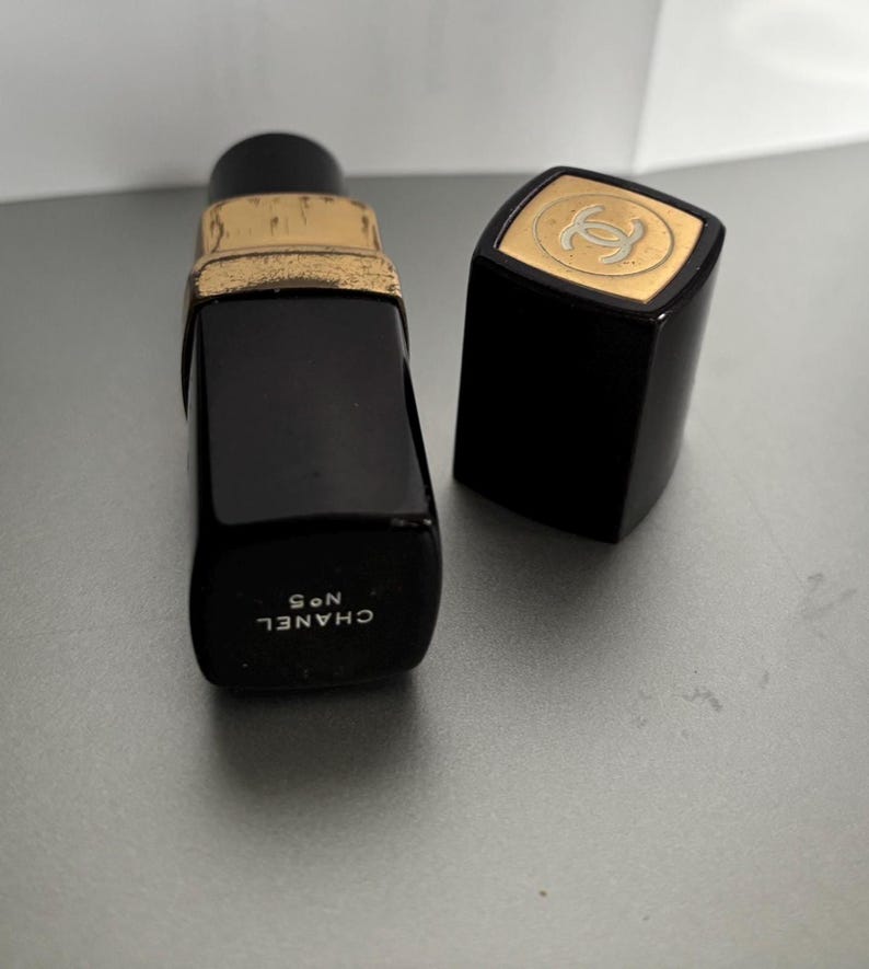 May include: A black perfume bottle with a gold-colored rim and a matching cap. The cap features a gold emblem. The bottle is labeled "CHANEL N&deg;5" in white text. The bottle is rectangular and has a classic design.