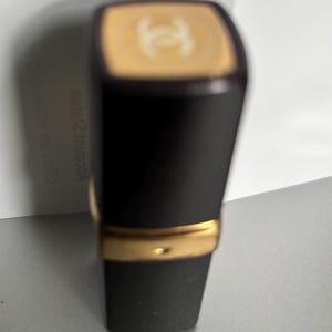 May include: Black lipstick tube with a gold band and a gold top featuring a logo. The lipstick has a matte black finish and a rectangular shape. The background is a light gray surface.
