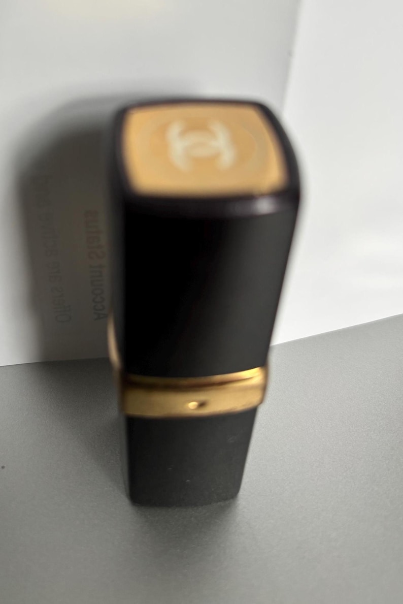 May include: Black lipstick tube with a gold band and a gold top featuring a logo. The lipstick has a matte black finish and a rectangular shape. The background is a light gray surface.
