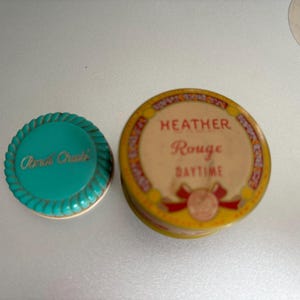 May include: Two vintage round compact cases. One is teal blue with a white rope design and the words "Avon Classic" in white. The other is yellow with a red ribbon and the words "Heather Rouge Daytime" in black.