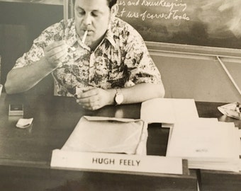 black and white photo of man at desk lighting cigarette and his name is hugh feely