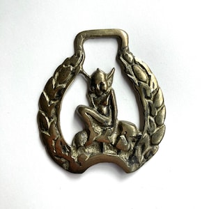 May include: A vintage metal horse brass featuring a seated fairy figure within a horseshoe-shaped frame. The frame is adorned with a laurel wreath design. The brass has a tarnished, antique finish, suggesting age and use. The fairy has pointed ears and is seated on a rock.